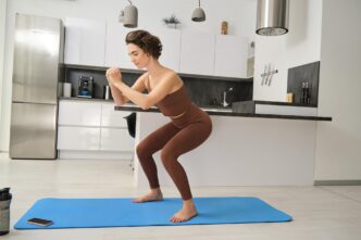 Young woman in activewear performs squats on a rubber mat during an indoor home workout in a brightly lit room.