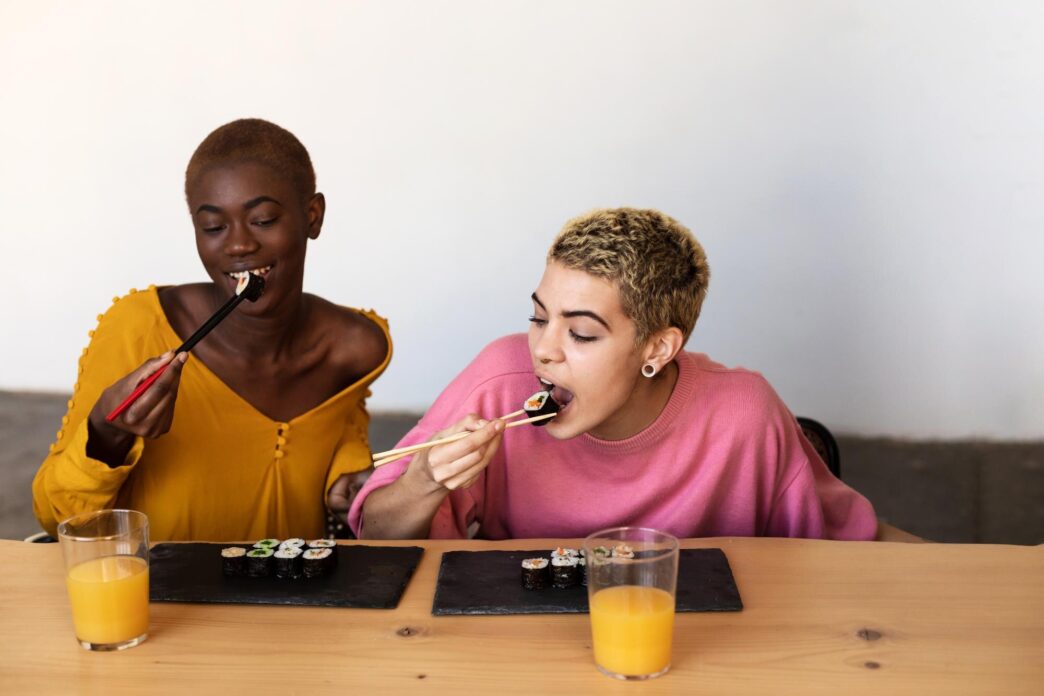 A lesbian couple smiles while eating sushi together in their home.