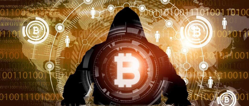 A hooded person uses a laptop to steal cryptocurrency, illustrating the concept of cyber theft.