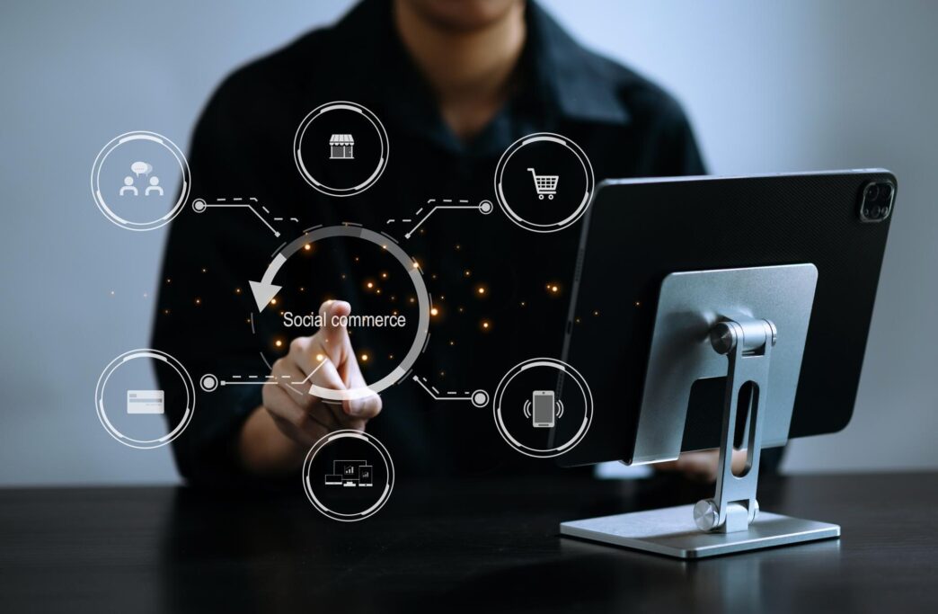 A man's hand uses a smartphone and tablet, displaying social commerce icons and illustrating social media marketing in an office setting.