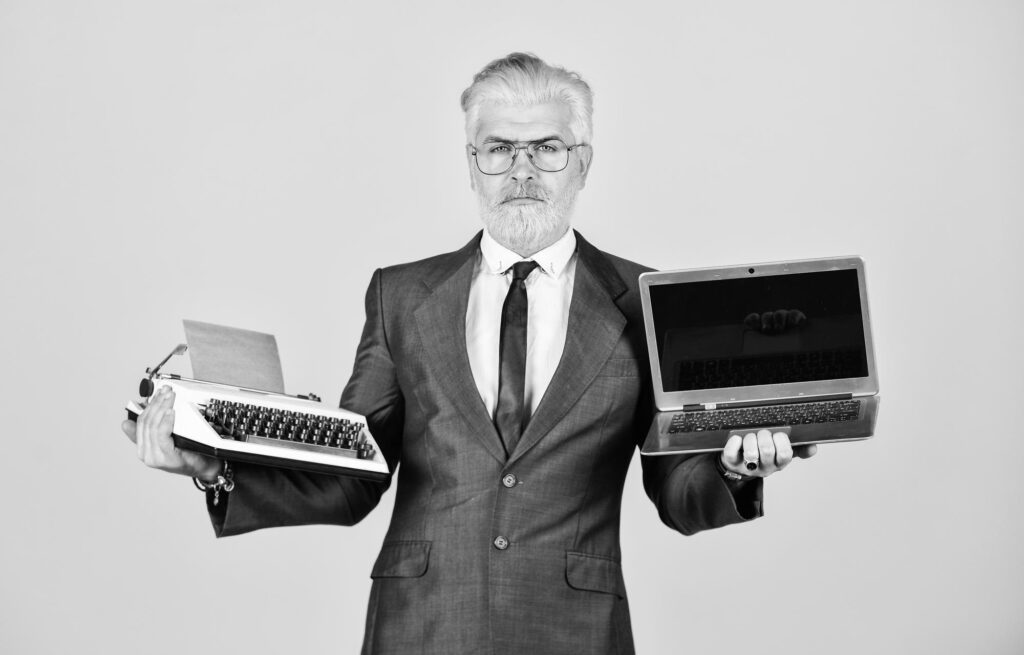 A mature man with a dyed beard uses both a vintage typewriter and a modern laptop, representing the intersection of old and new technology in business.