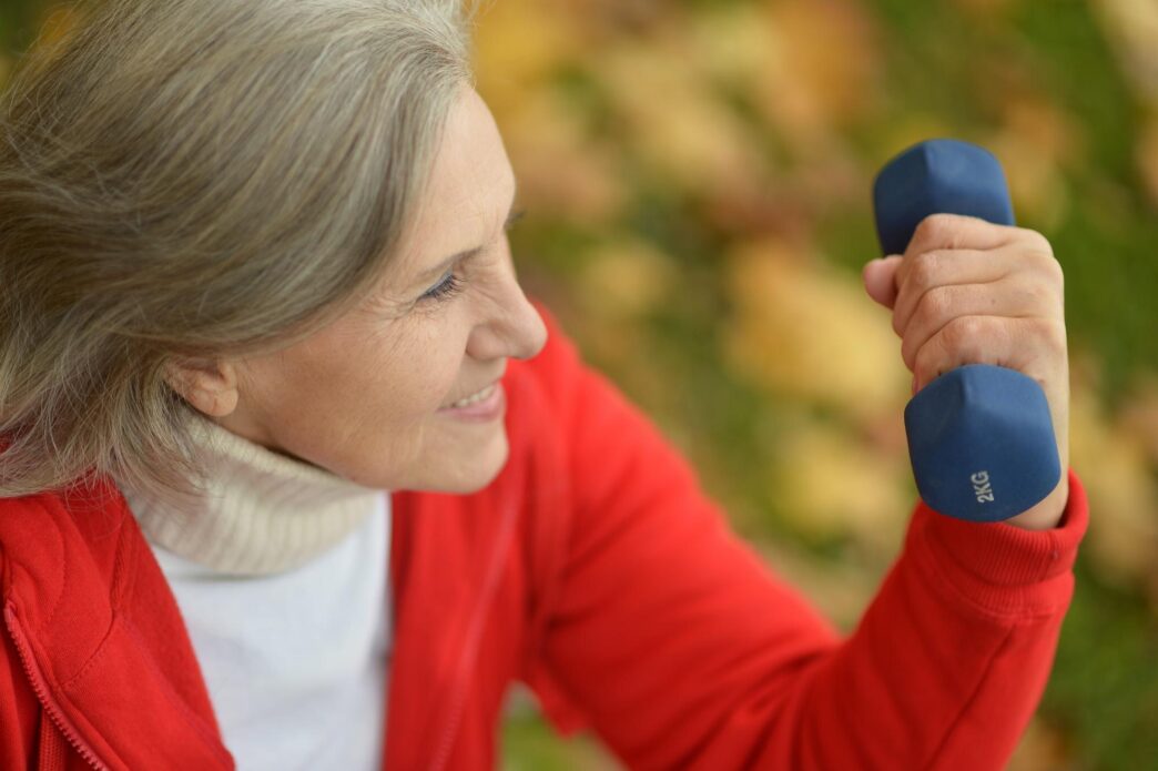 An older woman with gray hair lifts dumbbells while exercising in a park with fall foliage.