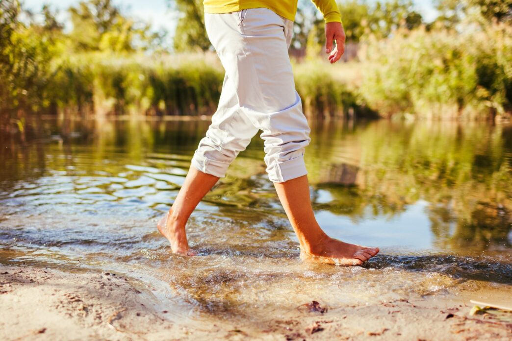 A middle-aged woman with bare feet walks along a riverbank in the autumn, with a close-up of her legs.
