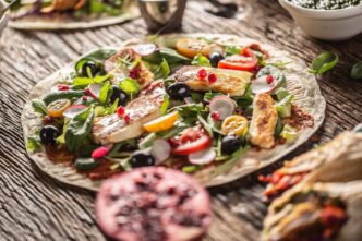 An overhead shot shows a vegetarian tortilla filled with salad, radishes, cherry tomatoes, olives, pomegranate seeds, and grilled halloumi cheese.