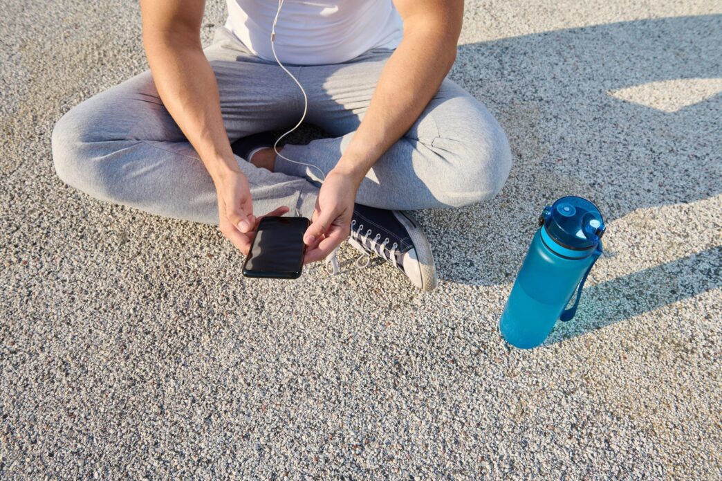 Athlete wearing earphones, sitting on a city bridge and checking their phone while relaxing after a morning cardio workout.