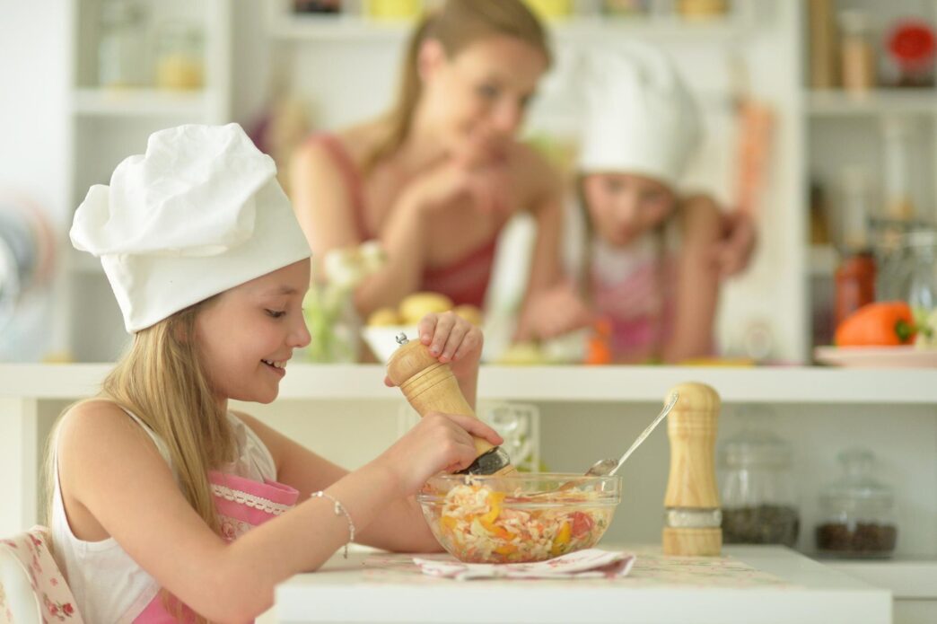 A young girl with a happy expression eats a salad in a kitchen.