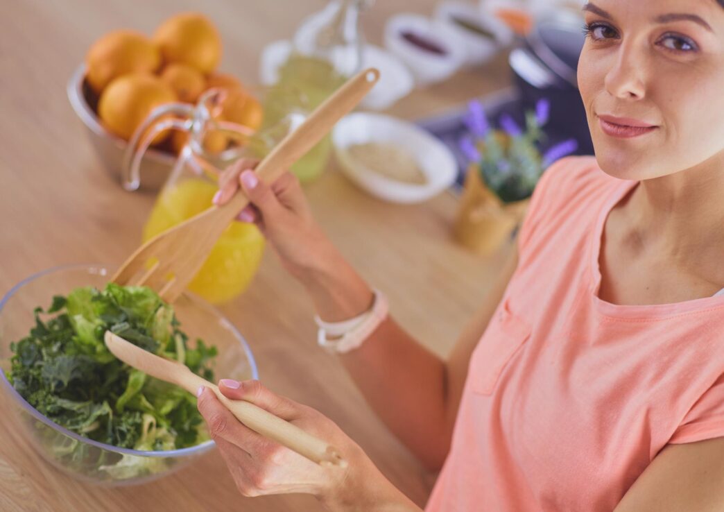 A young woman smiles while mixing a fresh salad in a kitchen.