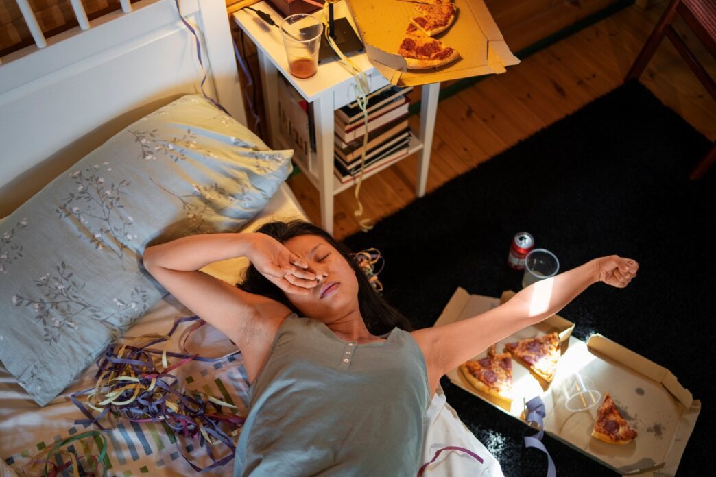 A top-down view shows a teenage girl lying in bed, surrounded by items like water bottles, tissues, and a phone, suggesting a hangover.