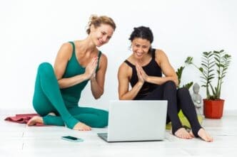 Two yoga instructors on a mat in a yoga studio smile and wave at the camera, welcoming viewers to their online yoga class.