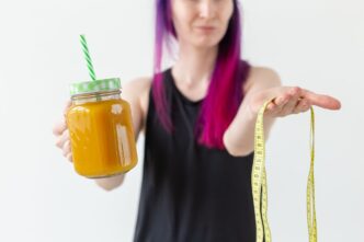 A young woman holds a fruity protein juice with a straw, with a measuring tape wrapped around her waist.