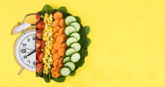 A white alarm clock and a plate of vegetables sit on a yellow surface, viewed from above with copy space.