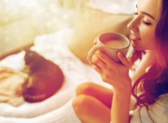 A smiling young woman in bed with a cat enjoys a warm beverage.