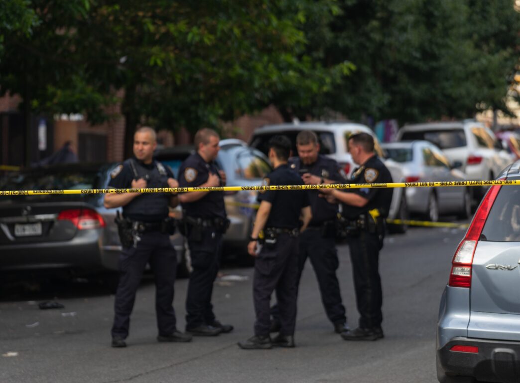 Five police officers stand on a taped-off street with parked cars during the day.