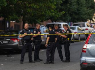Five police officers stand on a taped-off street with parked cars during the day.