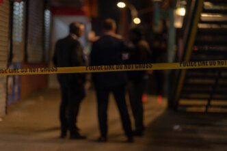 Nighttime crime scene with yellow police tape and three blurred detectives investigating outdoors.