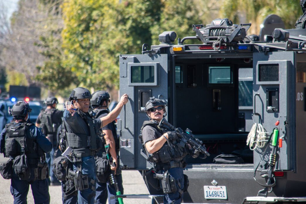 SWAT officers stand beside a gray Lenco armored vehicle, with one holding a rifle, ready for action.