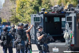 SWAT officers stand beside a gray Lenco armored vehicle, with one holding a rifle, ready for action.