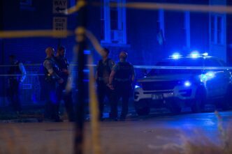 At night, few police officers stand behind yellow tape at a crime scene with flashing blue lights on a street.