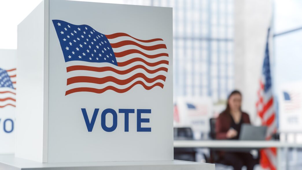 Voting booth with the American flag and "Vote" sign, person working at a table in the background, Elections_Gov.