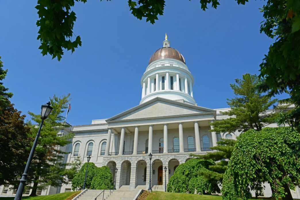 Maine State House with a prominent dome is located in Augusta, Maine, surrounded by greenery under a clear blue sky.