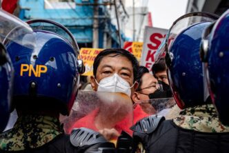A masked man peers through a clear shield, flanked by police in riot gear during a public demonstration.