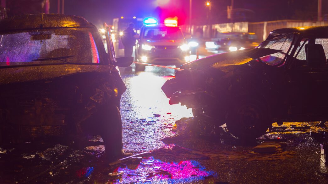 Nighttime car crash scene illuminated by police lights with damaged vehicles on a road.
