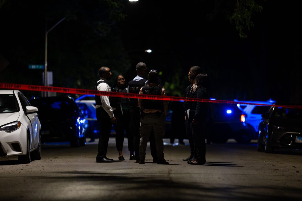 Police detectives converse at a nighttime homicide scene with flashing police car lights.