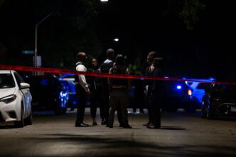 Police detectives converse at a nighttime homicide scene with flashing police car lights.