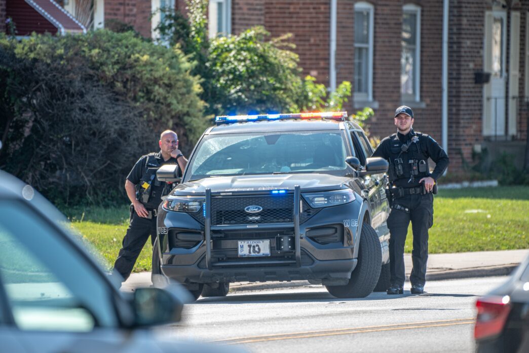 Two police officers stand by a black Ford Explorer with flashing lights on a residential street.