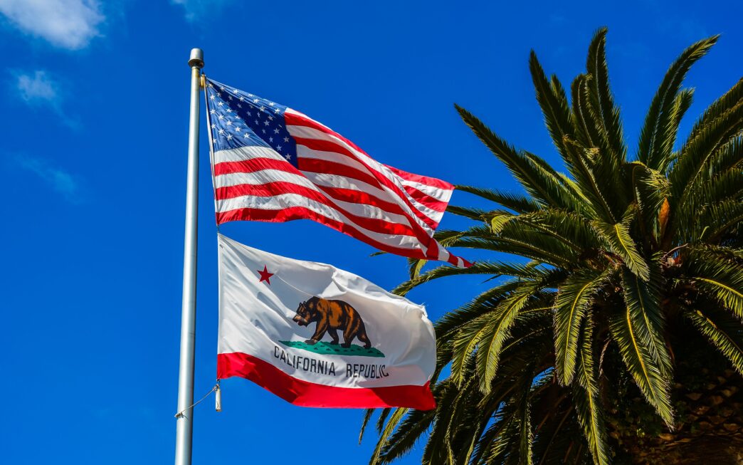 United States and California flags waving beside a palm tree under a clear blue sky.