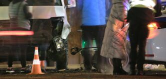 Heavily damaged cars on city street after collision, silhouettes of people present at night, highlighting road safety concerns.