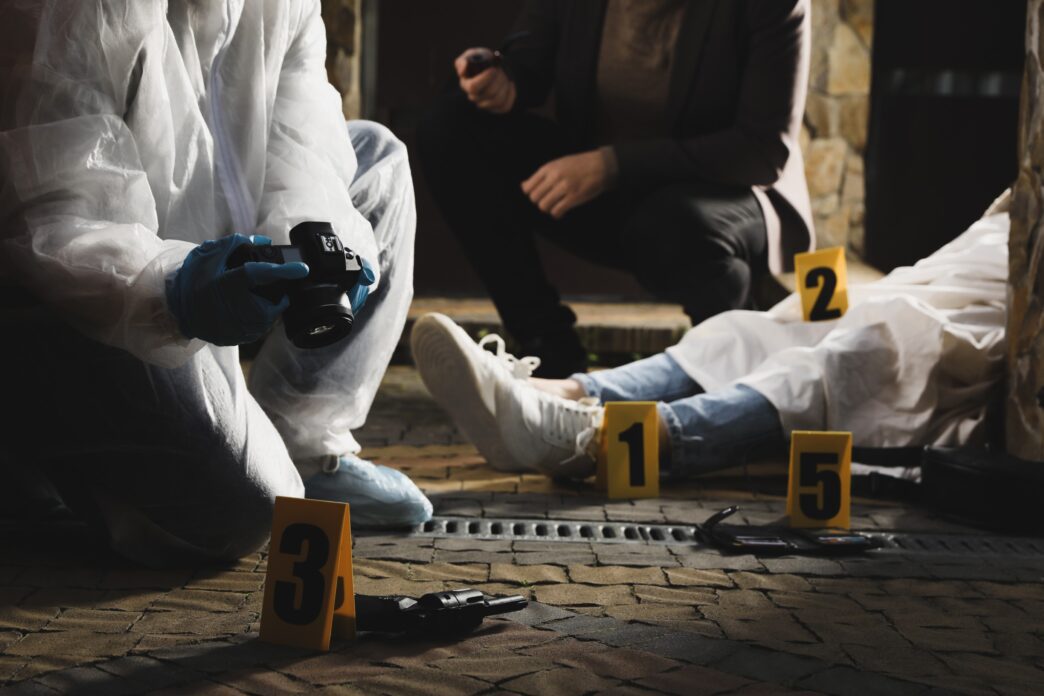 Investigator-criminologist examines outdoor crime scene with dead body and evidence markers.