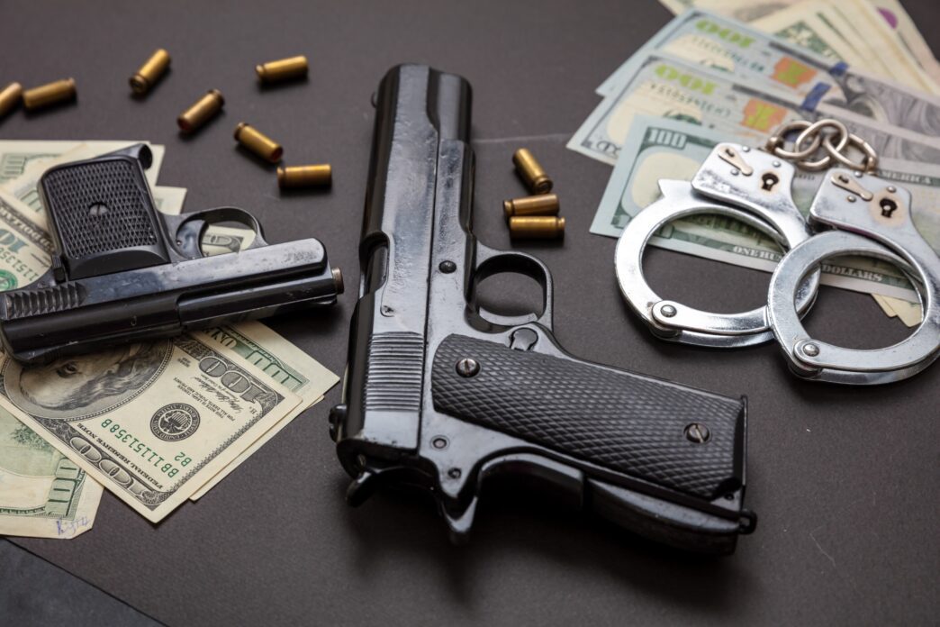 Pistol, handcuffs, and American dollar banknotes on a black background with bullet casings nearby.