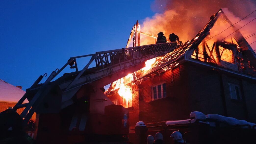 Firefighters extinguish a house fire in a village, using a crane amidst smoke and sparks.