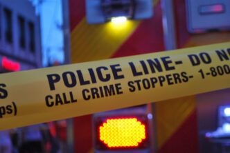 Yellow police tape reads "POLICE LINE-DO NOT CROSS CALL CRIME STOPPERS-1-800" against blurry emergency lights.