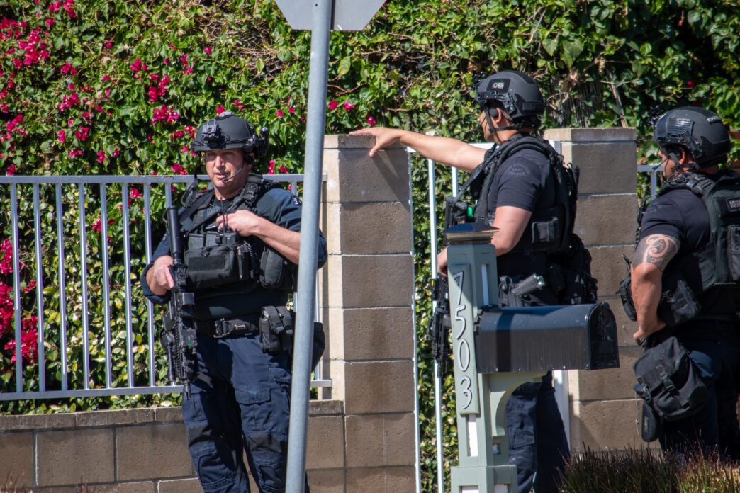 SWAT officers in tactical gear in suburb, one uses radio, another points at building for movement; Glenn Highcove-Shutterstock.