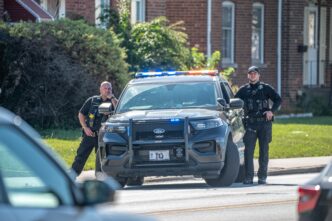 Two police officers stand by a black Ford Explorer with flashing lights on a residential street.