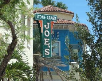 The green and orange "Joe's Stone Crab" sign partially visible between buildings and trees in Miami Beach.