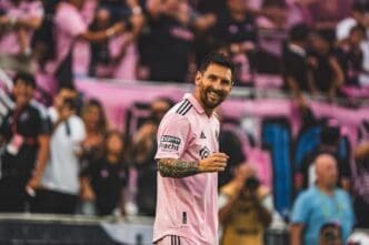 Lionel Messi, wearing an Inter Miami pink jersey, smiles broadly and looks towards the camera on a soccer field, with blurred fans in pink and black behind him.