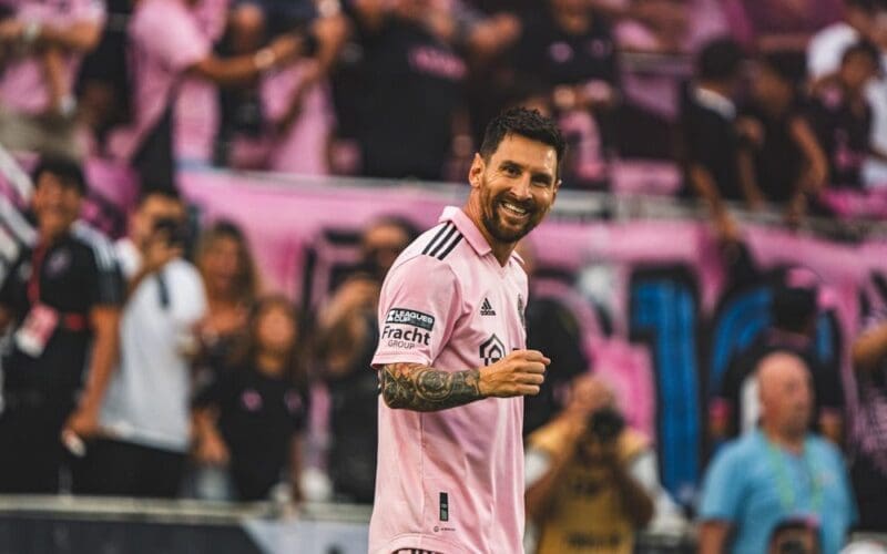 Lionel Messi, wearing an Inter Miami pink jersey, smiles broadly and looks towards the camera on a soccer field, with blurred fans in pink and black behind him.