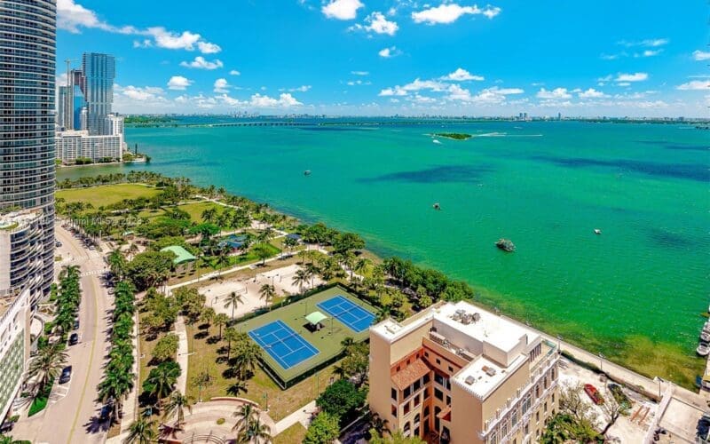 An aerial view of Margaret Pace Park in Miami, showcasing its green lawns, palm trees, and tennis courts, situated along the edge of a large bay with turquoise water, a bridge, and a city skyline in the distance under a bright blue sky.
