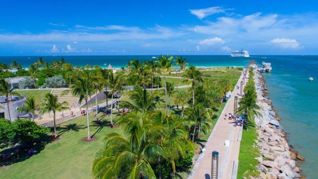 An aerial view of South Pointe Park in Miami Beach, showing lush green lawns, palm trees, pathways, a pier, and the ocean with a cruise ship.