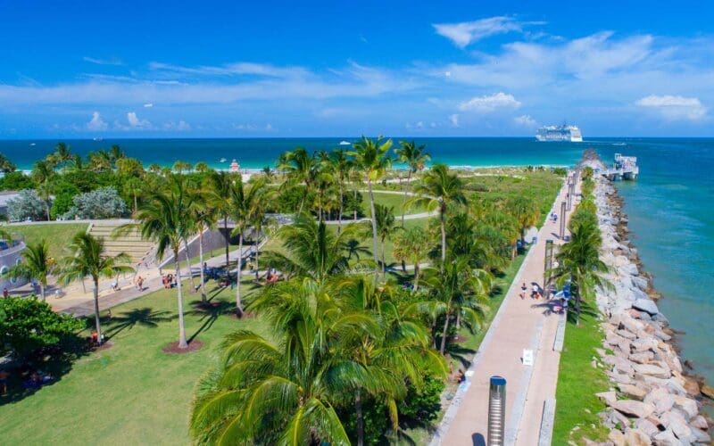 An aerial view of South Pointe Park in Miami Beach, showing lush green lawns, palm trees, pathways, a pier, and the ocean with a cruise ship.