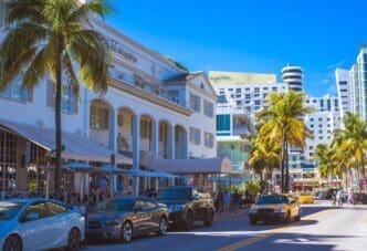 The elegant white facade of The Betsy Hotel, an Art Deco building with a colonnade, stands on a busy Ocean Drive. Parked cars, palm trees, outdoor seating, and other modern and classic buildings are in the background under a clear blue sky.