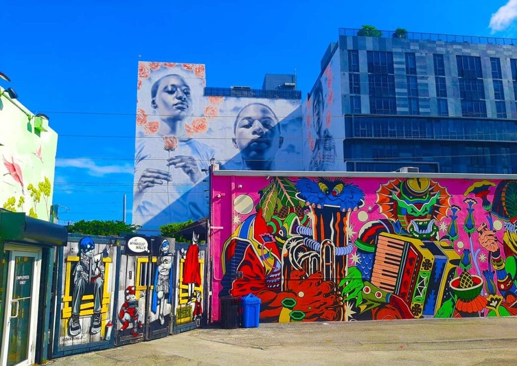 A vibrant street scene in Wynwood, Miami, featuring multiple large-scale murals: a pink wall with colorful abstract and figurative art, and a building facade with grayscale portraits surrounded by roses, all under a clear blue sky.