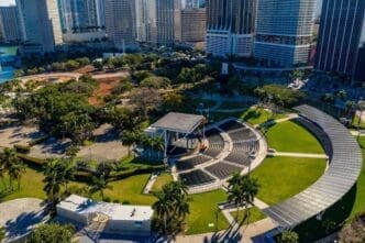 An aerial view of the Bayfront Park Amphitheater in Miami, showing its semi-circular seating, stage with a roof, and surrounding green parkland and palm trees, with a dense city skyline in the background.